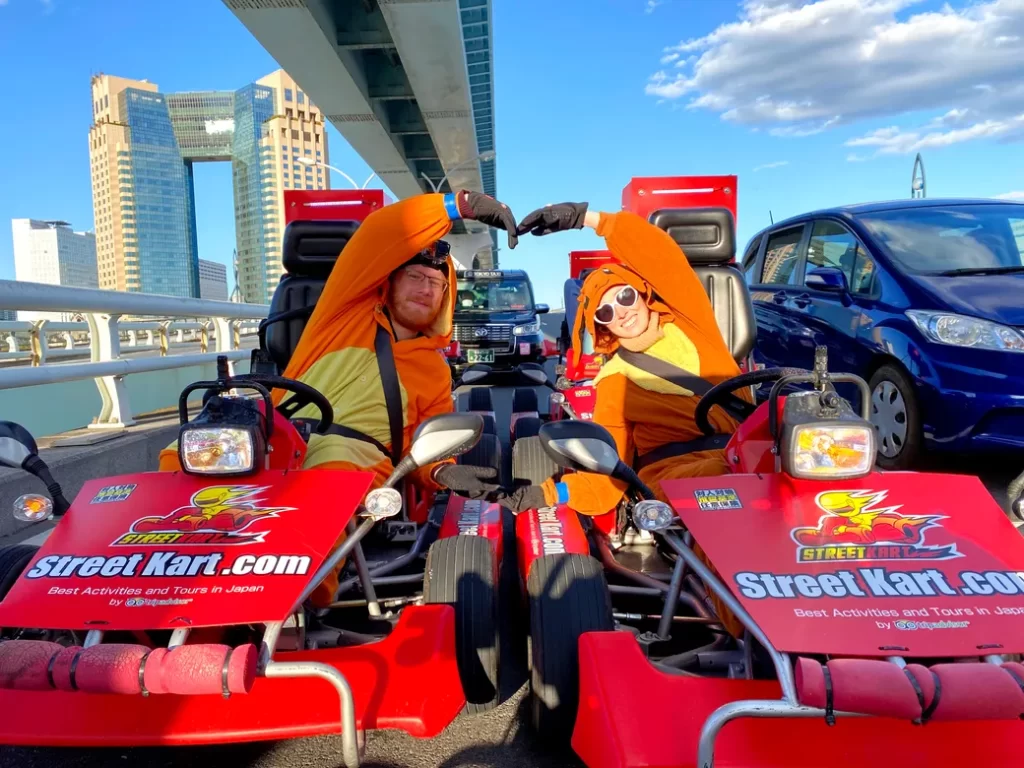 Two people in orange costumes form a heart with their arms while sitting in red go-karts on a sunny bridge, with modern buildings in the background and a blue sky.