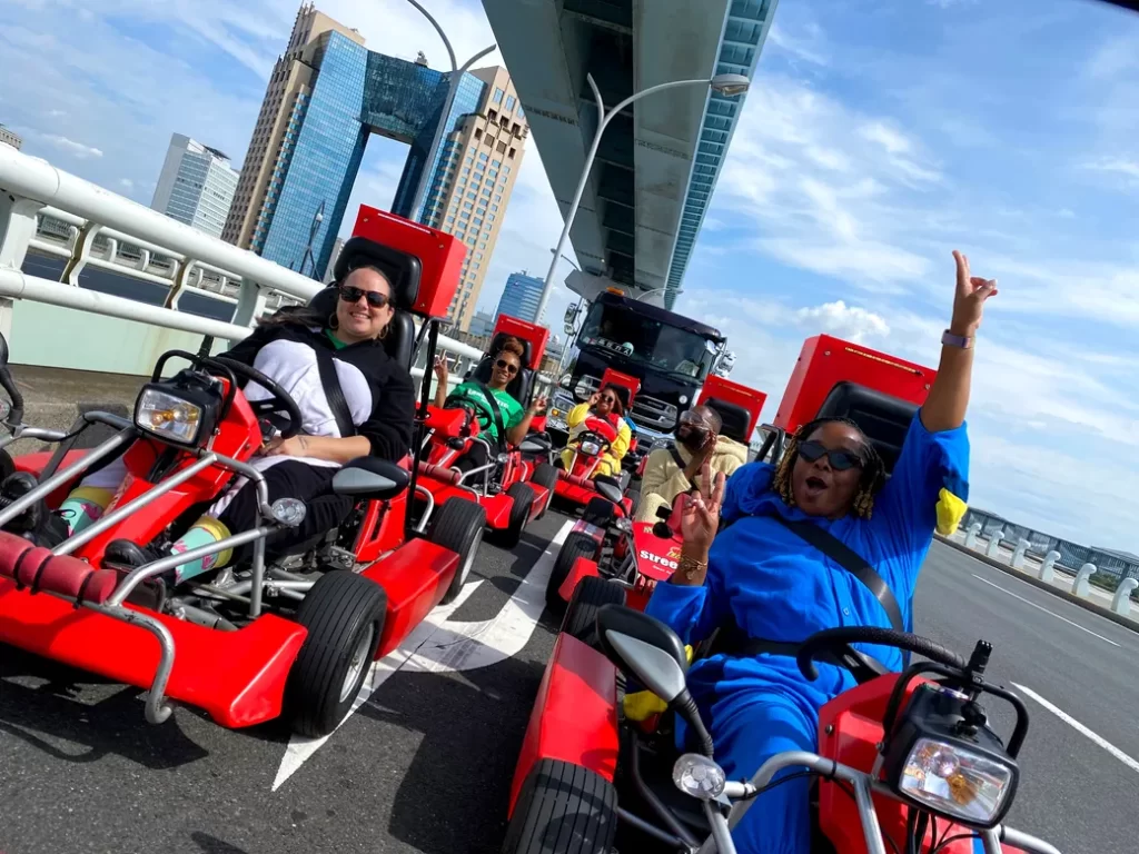 IMG_4156 - StreetKart Group of people riding red go-karts on a bridge, city skyline in the background, woman in blue raising her hand cheers at the front.